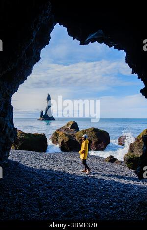 Eine Frau, die in einer felsigen Höhle steht, bewundert den atemberaubenden Strand von Reynisfjara. Majestätisch erheben sich die dramatischen Meeresstapel im Hintergrund, während die Wellen in Island gegen die Küste krachen Stockfoto