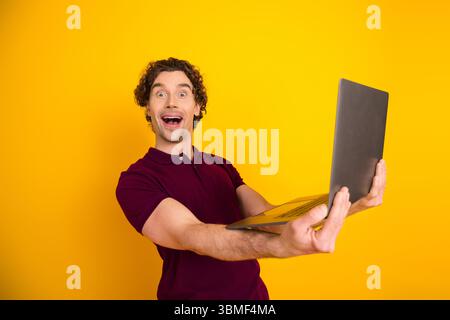 Aufgeregter junger Mann, der ein Notebook auf leuchtendem gelbem Hintergrund hält und Glück, Technologie und modernen Lebensstil zeigt Stockfoto