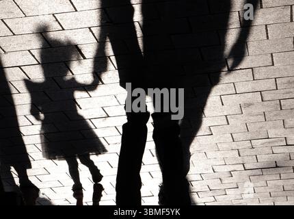 Schatten-Silhouette eines Kleinkindes und ihrer Eltern, die Hand in Hand auf der Sommerpromenade gehen, in Schwarz-weiß mit hohem Kontrast Stockfoto