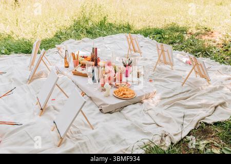 Picknick im Freien mit leeren Leinwänden, Kunstbedarf, Getränken und Snacks auf weichem weißem Stoff auf einer sonnigen Wiese. Stockfoto