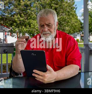 Ein älterer Mann mit Bart sitzt an einem Tisch auf einer Veranda. Er trägt ein rotes Hemd und schaut sich ein Tablet an. Er wirkt engagiert und zufrieden, während er die Sonne genießt Stockfoto