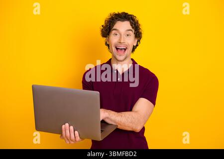 Ein aufgeregter junger Mann hält einen Laptop mit fröhlichem Ausdruck vor einem leuchtend gelben Hintergrund, der Freude zeigt Stockfoto