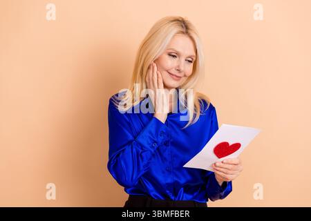 Elegante Frau in einer leuchtend blauen Bluse mit einem herzhaften Buchstaben und einem roten Herzemblem, umgeben von einem weichen beigefarbenen Hintergrund. Stockfoto