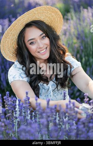 Junge schöne Frau mit langen dunklen Haaren in einem Hut, die Lavendelblumen auf einem Feld pflückt. Hält ein Glas ätherischen Öls in ihren Händen. Stockfoto