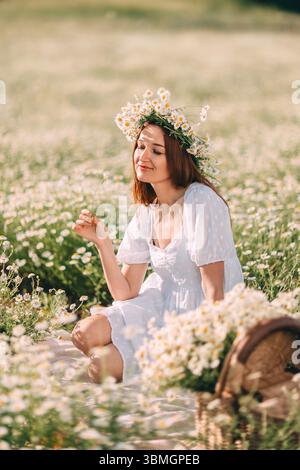 Gänseblümchen Frau Sommer: Frau sitzt am Sommertag im Gänseblümchenfeld und hält Blumen, Korb in der Nähe für Picknickfreude. Stockfoto