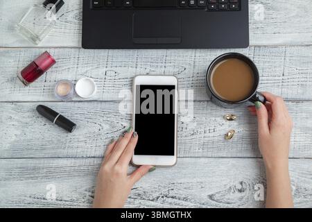 Schreibtisch. Frauenarbeitsplatz. Damenhände, Smartphone, Laptop, Kaffeetasse, Parfüm und Kosmetik. Die Aussicht von oben. Flache Lagen Stockfoto