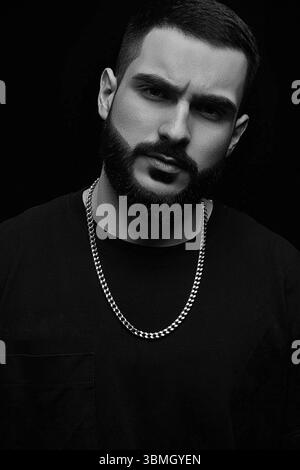 Nahaufnahme eines dramatischen Porträts eines jungen ernsthaften Mannes, Musikers, Sängers, Rappers mit einem Bart in schwarzer Kleidung auf einem schwarzen isolierten Hintergrund. Schwarz A Stockfoto