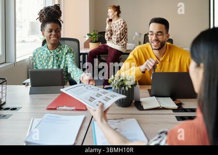 Junge Schwarze Frau und Mann arbeiten am Coworking Tisch mit digitalen Tablets zusammen, während Kaukasier am Telefon spricht, asiatische Frau Dokumente überreicht Stockfoto