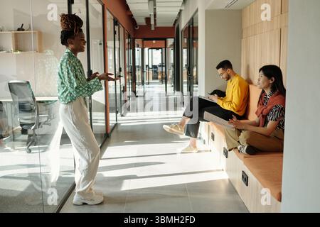 Junge Schwarze Frau, die im modernen Korridor des Coworking-Büros auf einer Bank steht und Gesten macht, während sie mit einem jungen, rassistischen Mann und einer jungen asiatischen Frau spricht Stockfoto
