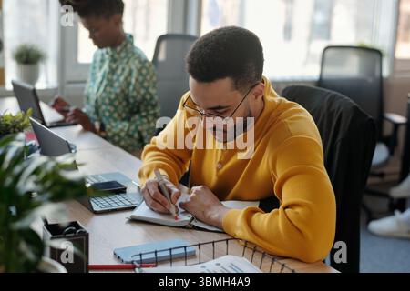 Junger Schwarzer Mann mit gelbem Pullover, der in Notizbuch am gemeinsamen Schreibtisch schreibt, während junge Erwachsene Schwarze Frau in gemustertem Hemd am Laptop arbeitet Stockfoto