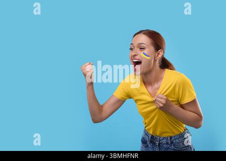 Aufgeregter Fan mit Flagge der Ukraine auf ihrer Wange vor hellblauem Hintergrund. Leerzeichen für Text Stockfoto