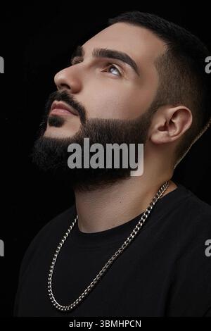 Ein dramatisches Porträt eines jungen seriösen Mannes, Musikers, Sängers, Rappers mit einem Bart in schwarzer Kleidung auf einem schwarzen isolierten Hintergrund. Er schaut hoch Stockfoto