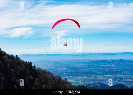 Hoch über einem üppigen Tal gleitet ein Gleitschirmflieger mit einem leuchtenden roten Baldachin anmutig durch den Himmel, umgeben von majestätischen Bergen und atemberaubend Stockfoto