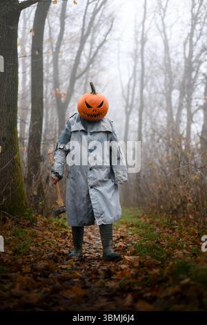 Vogelscheuche mit Axt in einem alten zerrissenen Mantel und Kürbiskopf aus Laterne sorgen für Horror-Atmosphäre. Gruselige Halloween-Figur in verwunschener Waldlandschaft Stockfoto
