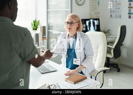 Kaukasische Ärztin mittleren Alters sitzt am Schreibtisch und berät Schwarzer Mann mit Diabetes in der medizinischen Praxis, Patient übergibt Klemmbrett an Arzt, Gluc Stockfoto