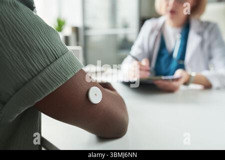 Schwarzer junger erwachsener Mann, der Glukosemonitor am Oberarm trägt, Beratung mit kaukasischer Ärztin mittleren Alters in der Arztpraxis, Doktor schreibt Notizen Stockfoto