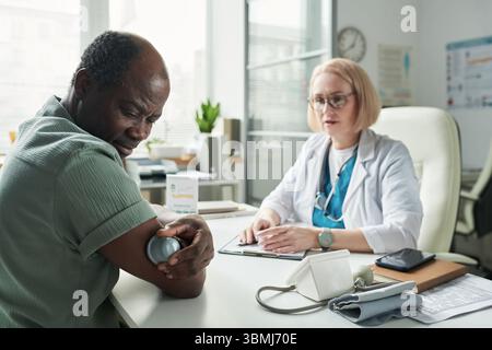 Schwarzer Mann mittleren Alters, der Blutzucker mit Glucometer überprüft, während er gegenüber von der kaukasischen Ärztin mittleren Alters in der Arztpraxis während der di sitzt Stockfoto