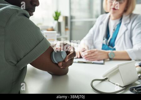 Schwarzer Mann mittleren Alters, der den Blutzucker mit einem medizinischen Gerät überprüft, während er gegenüber von kaukasischer Ärztin mittleren Alters in der Arztpraxis DURIN sitzt Stockfoto