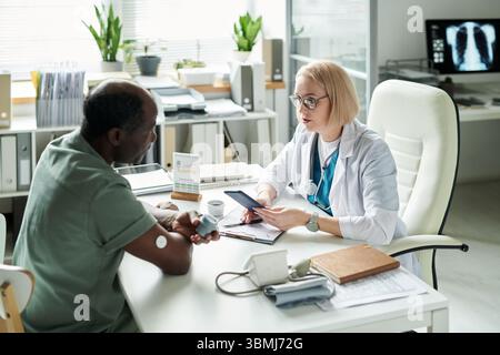 Schwarzer Mann mittleren Alters, der gegenüber von der kaukasischen Ärztin sitzt, die Diabetes-Management während der medizinischen Beratung in einem modernen Büro diskutiert, Blut g Stockfoto