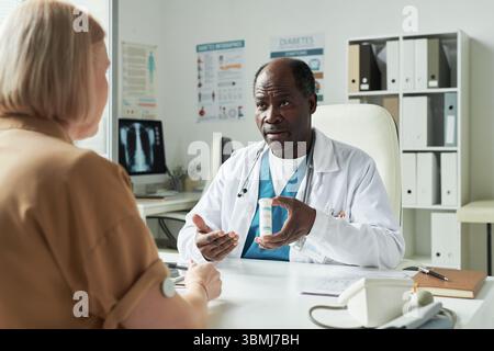 Schwarzer männlicher Arzt mittleren Alters, der Medikamente für Patienten der kaukasischen Frau mittleren Alters während der Diabeteskonsultation in der Arztpraxis erklärt, verschreibungspflichtig Stockfoto