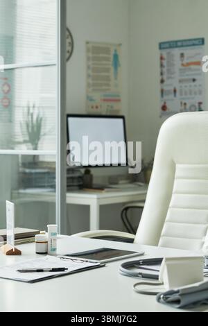 Leeres medizinisches Büro mit Schreibtisch mit Blutdruckmessgerät, verschreibungspflichtigen Flaschen, Klemmbrett mit Dokumenten, Computermonitor und Poster an der Wand, r Stockfoto