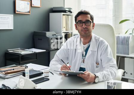 Porträt eines männlichen kaukasischen Arztes mittleren Alters, der am Schreibtisch sitzt, Klemmbrett und Stift hält und in die Kamera in der Arztpraxis während der Konsultation mit schaut Stockfoto