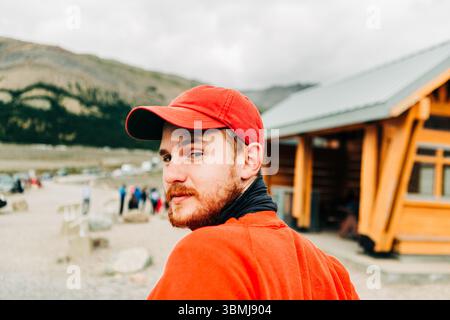 Ein Mann mit rotem Hut und Hemd schaut in die Nähe der Lodge zurück, in Berglandschaft. Stockfoto
