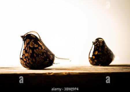 Zwei handgefertigte Vogelfiguren aus Glas, eine in scharfem Fokus, mit dunklen und goldenen gesprenkelten Mustern, stehen auf einer rustikalen Holzoberfläche. Stockfoto