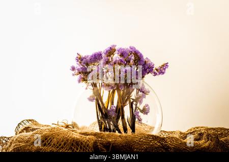 Eine Vase aus klarem Glas, gefüllt mit wunderschönen lila getrockneten Blumen, elegant auf einem rustikalen Jutestoff vor einem weichen, hellen Hintergrund angeordnet. Stockfoto