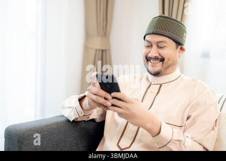 Junger muslimischer Mann, der zu Hause auf dem Sofa sitzt und in entspannter Atmosphäre lächelt, während er das Smartphone benutzt. Stockfoto