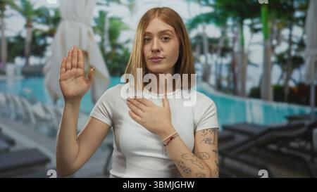Junge Frau mit Tätowierungen am Pool im Resort, Hand auf Brust in ruhiger Umgebung im Freien mit Stühlen und Palmen Stockfoto