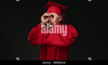 Junger chinesischer Mann in roter Graduierungsmütze und rotem Kleid macht binokulare Gesten isoliert auf schwarzem Hintergrund, die Neugier und Zukunftsvision symbolisieren. Stockfoto