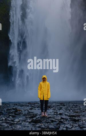Eine Frau, die vor dem majestätischen Skogafoss-Wasserfall steht, ist eine Reisende in einer leuchtenden gelben Regenjacke umgeben von Nebel und atemberaubender natürlicher Schönheit. Stockfoto