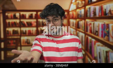 Junger bärtiger hispanischer Mann mit Brille und gestreiftem Hemd steht selbstbewusst in einer Bibliothek, umgeben von Regalen mit bunten Büchern, die eine intel-information erschaffen Stockfoto