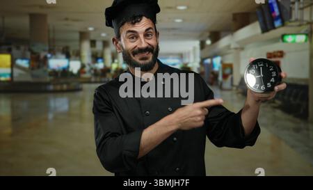 Der hispanische Koch in schwarzer Uniform zeigt auf die Uhr im Flughafenterminal und unterstreicht die Dringlichkeit der Zeit in der geschäftigen Reiseumgebung. Stockfoto