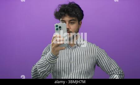 Junger Mann mit Bart, der das Smartphone vor einem leuchtend violetten Hintergrund hält, während er ein gestreiftes Hemd trägt und sich auf sein Gerät konzentriert, möglicherweise beim SMS oder Stockfoto