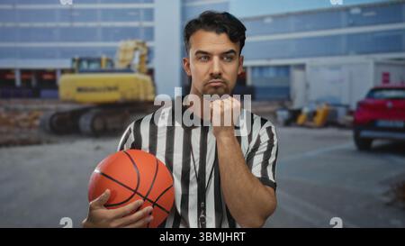 Junger hispanischer Mann, der Basketball auf einer Baustelle in Schiedsrichteroutfit hält nachdenklicher Ausdruck Outdoor-Umgebung städtische Umgebung Stockfoto