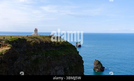 Besucher bewundern die atemberaubende Aussicht von Dyrholaey, einem prominenten Vorgebirge in Island. Der legendäre Leuchtturm steht stolz vor der Kulisse des endlosen Ozeans und der zerklüfteten Klippen. Stockfoto