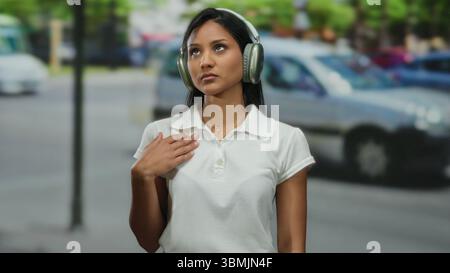 Eine Frau mit Kopfhörern steht auf einer belebten Straße mit vorbeifahrenden Autos, absorbiert in Gedanken, jugendlicher Energie und Konzentration, die in ihrem Verhalten erkennbar ist. Stockfoto