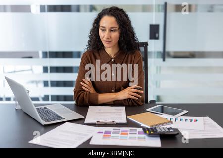 Eine Geschäftsfrau sitzt tief in Gedanken an ihrem Schreibtisch, umgeben von Dokumenten und einem Laptop, in einem modernen Büro. Stockfoto