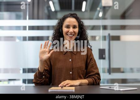 Eine lächelnde Frau mit lockigen Haaren, die an der Kamera schwingt, während sie im Büro ein Headset trägt. Stockfoto
