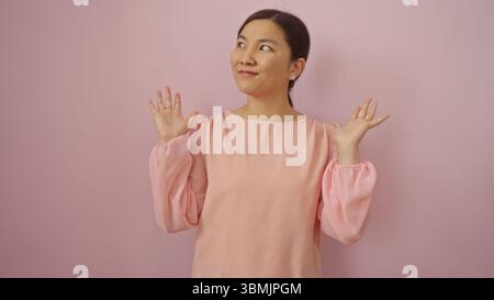 Frau lächelt in rosafarbener Bluse vor pastellrosa Hintergrund und zeigt Freude und Charme mit erhobenen Händen, verkörpert Anmut und Eleganz. Stockfoto