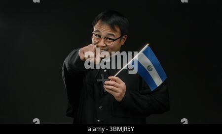 Priester mit salvadorianischer Flagge vor dunklem Hintergrund mit selbstbewusstem Ausdruck und Kreuzkette, die Glauben und multikulturelle Identität symbolisiert. Stockfoto