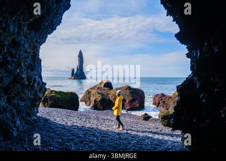 Ein atemberaubender Blick auf Reynisfjara Beach mit hoch aufragenden Basaltsäulen und Meereswellen. Ein Besucher in einer gelben Jacke erkundet die einzigartige Landschaft und fängt die Essenz der natürlichen Schönheit Islands ein. Stockfoto