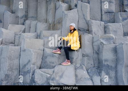Ein Reisender sitzt auf den majestätischen Basaltsäulen des Reynisfjara Beach, Island. Die einzigartige Felsformation trägt zur atemberaubenden Küstenlandschaft unter klarem Himmel bei und schafft ein unvergessliches Erlebnis. Stockfoto