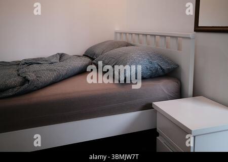 Seitlicher Blick auf ein ungemachtes Bett mit geometrisch gemusterten Kissen und Federdecke in einem gemütlichen weißen Schlafzimmer. Gemütliches Schlafzimmer in einem Landhaus oder Hotel Stockfoto