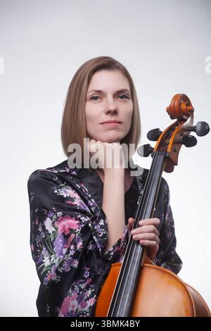 Erwachsene kaukasische Frau in schwarzem Blumengewand, die ein Cello in einem Atelierporträt hält, die Hand auf dem Kinn ruht. Stockfoto