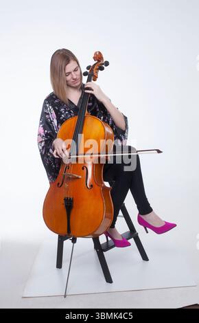 Mitte der 20er bis 30er Jahre sitzt kaukasische Frau in schwarzem Blumengewand, schwarzer Hose und rosafarbenen Heels in einem Studio mit weißem Hintergrund, das Cello spielt. Stockfoto