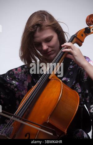 Junge Erwachsene kaukasische Frau spielt Cello im Studio, trägt ein schwarzes Blumengewand, fokussiert mit den Augen nach unten. Stockfoto