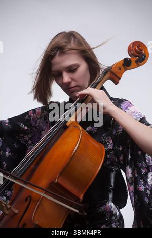 Junge Erwachsene Kaukasierin, Cellospielerin, konzentriert sich auf das Instrumentenspielen im Studio. Ich trage einen schwarzen Kimono-Mantel mit Blumenmuster. Stockfoto